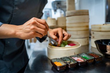 Bambu buharlı bir tencerenin içindeki hamur köftelerine malzeme ekleyen bir şefin yakın plan fotoğrafı, çeşitli baharatlar ve malzemeler paslanmaz çelik tezgahın üzerine dizilmiş.
