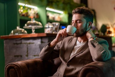 Spor ceketli, rahat bir kafede kahve içen, rahat ve sofistike bir ortamda rahatlığı vurgulayan profesyonel bir adam..