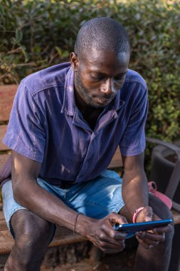 Genç siyahi bir adam parktaki ahşap bir bankta oturuyor, akıllı telefonuna konsantre bir şekilde bakıyor. Gündelik yaz kıyafeti, doğal ışık ve rahat duruş dijital odaklanma anı olduğunu gösteriyor..