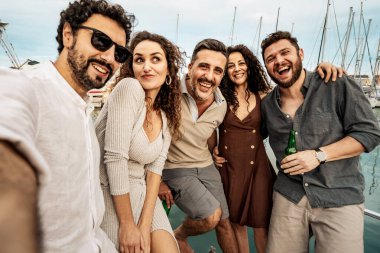 Bir grup neşeli arkadaş marinadaki yat partisinde komik bir selfie çekiyorlar. Deniz kenarında gülüyorlar ve yaz akşamının tadını çıkarıyorlar..