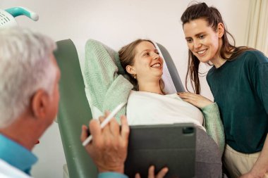 Lezbiyen bir çift, bir doktorla doğum öncesi muayeneye katılıyor. Heyecanlı bir şekilde bebeklerini bekliyorlar. Görüntü, LGBTQ + ebeveynlik, tıbbi destek ve modern bir klinikte hamileliğin neşesini vurguluyor.