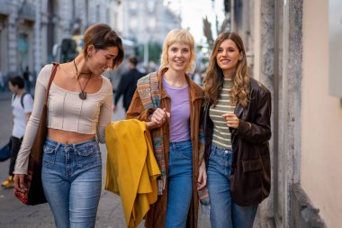Üç modaya uygun genç bayandan oluşan bir grup neşeli bir şehir caddesinde yürüyor, gülümsüyor ve gülüyor. Neşeli ifadeleri, modaya uygun kıyafetleri ve günlük etkileşimleri güçlü bir arkadaşlığı vurguluyor..