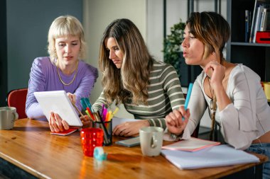 Modern bir ofiste iş birliği yapan üç genç yaratıcı kadın dijital tablet kullanarak fikirleri tartışıyor ve not alıyorlar. Profesyonel bir ortamda takım çalışması ve yenilik.