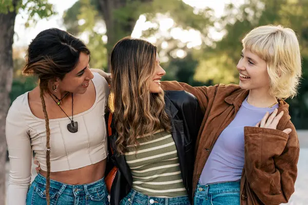 Canlı bir sohbete tutulan üç genç kadın, sıradan bir açık hava ortamında birlikte gülüyorlar. Şık kıyafetleri ve neşeli ifadeleri dostluğu ve bağlantıyı vurguluyor..