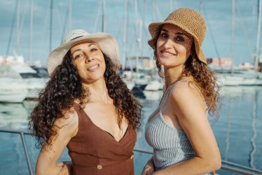 İki neşeli kadın yaz şapkası takıyor ve marinada gülümsüyor, deniz kenarında güneşli bir tatil gününün tadını çıkarıyorlar. Yaz seyahati ve arkadaşlık için mükemmel bir kavram..