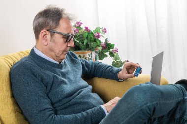 Sarı bir koltukta oturan yaşlı bir adam, bir kredi kartı ve dizüstü bilgisayarla online alışveriş yapıyor. Dijital finansman ve e-ticaret güvenliğine odaklı ev ortamı.