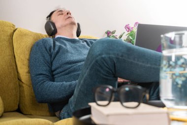 Orta yaşlı bir adam sarı bir koltukta dinleniyor, gürültüyü önleyen kulaklıklarla müzik dinliyor. Ev ortamında bir dizüstü bilgisayar, kitaplar ve bardaklar görülebilir..
