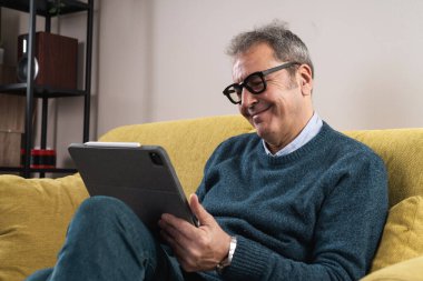 Bir tablet kullanarak kanepede oturan mutlu kıdemli bir adam, evde dijital içeriği izlerken ya da gezinirken gülümsüyor. Modern teknoloji, bağlantı ve rahatlama kavramı.
