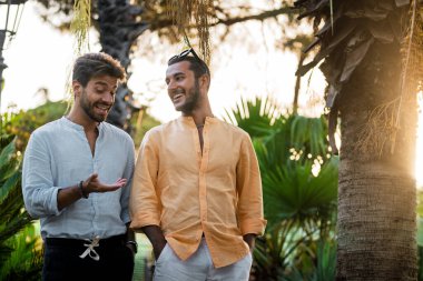 İki şık genç adam gün batımında tropikal bir parkta neşeli bir şekilde sohbet ediyor, arkadaşlığın ve bağlantının keyfini çıkarıyorlar..