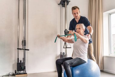 Rehabilitasyon terapisi sırasında fizyoterapist desteğiyle direnç bandı egzersizi yapan yaşlı bir kadın..