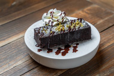 Taze krem şanti ve çikolata yağmuruyla kaplı zengin ve nemli çikolatalı browni, ahşap bir masada beyaz bir tabakta servis edilir..