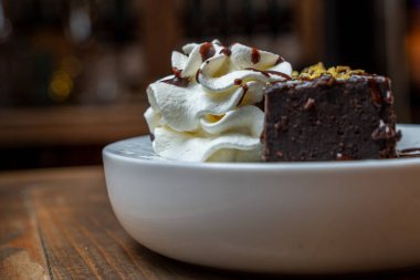 Çikolatalı browni diliminin yakın plan fotoğrafı, krem şantili, çikolata soslu, sıcak bir restoranda beyaz bir tabakta servis edilen.