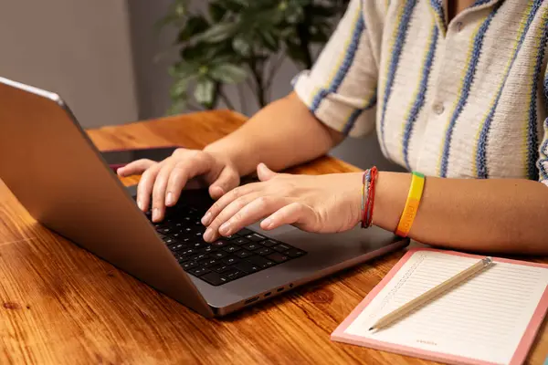 Not defteri ve gurur bilekliğiyle dizüstü bilgisayarda yazı yazan ellerin yakın plan, uzaktan çalışma, yaratıcılık ve kapsayıcılığı temsil ediyor..