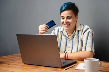 Evde bilgisayarını kullanarak internetten alışveriş yaparken elinde kredi kartı tutan mavi saçlı gülümseyen genç bir kadın. Dijital ödemeler ve modern e-ticaret kavramı.