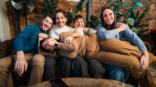 Genç, çeşitli arkadaşlar, arkadaşlarını kanepenin karşısında kucaklıyor, gülümsüyor ve neşeli bir enerjiyle kapalı mekanda eğleniyorlar..