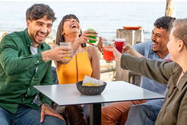 Bir grup farklı genç arkadaş deniz kenarındaki bir kafede renkli içkilerle gülüp kadeh kaldırarak neşeli ve rahat bir atmosferin tadını çıkarıyorlar..