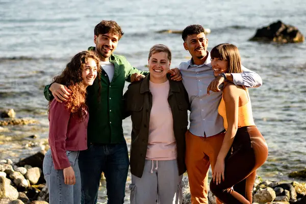 Güneşli bir günde, deniz kenarında gülümseyen ve kaynaşan bir grup arkadaş birlikte neşeli ve rahat bir anın tadını çıkarıyorlar..