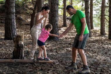 Orman macerası sırasında kızın kütüğün üzerinde yürümesine yardım eden ebeveynler, doğada güven, denge ve aile bağını sembolize ediyor..