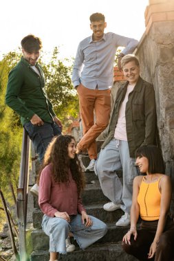 Çeşitli arkadaş grupları, doğal ışıkta gülüp kaynaşarak, açık hava basamaklarında gündelik sohbetin keyfini çıkarıyorlar..