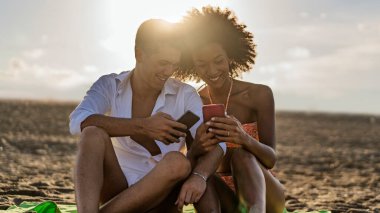 Gün batımında kumsalda oturan neşeli genç çift akıllı telefonlarına bakıp romantik bir yaz anının tadını çıkarırken gülümsüyor..