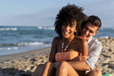 Yaz tatili boyunca kumsalda oturup, sevgi ve günışığının tadını çıkarırken gülümseyen genç bir çift..