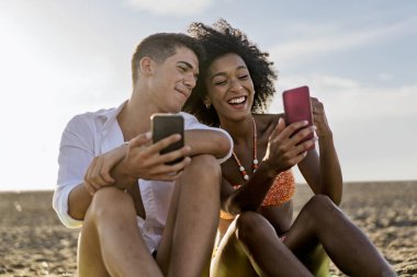Neşeli genç çift güneşli bir sahilde birlikte oturup akıllı telefonlar kullanıyor, yaz tatili boyunca gülüyor ve kaynaşıyorlar..