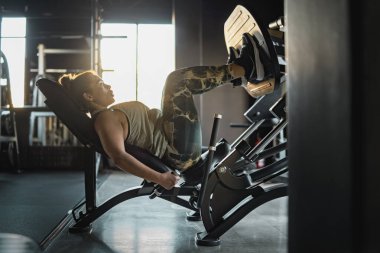 Spor salonunda egzersiz yaparken bacak presi makinesinde çalışan kendinden emin genç bir kadın. Güç, zindelik ve azim kavramı.