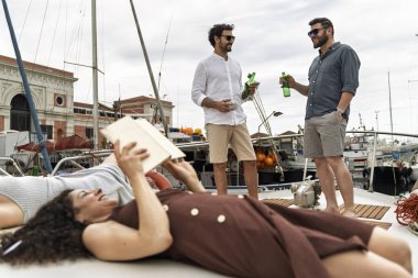 Tekne güvertesinde bir grup arkadaş marinada dinlendirici bir yaz gününde içki içip kitap okuyorlar.