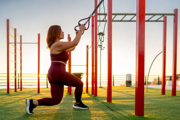Gün batımında açık hava spor salonunda direniş gruplarıyla çalışan atletik bir kadın fitness, güç ve sağlıklı bir yaşam tarzını teşvik ediyor..
