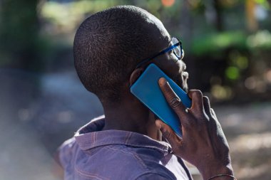 Gözlüklü bir adamın doğal ışık altında akıllı telefonda konuşması, günlük kıyafetler ve sığ alan derinliği olan arka plan.