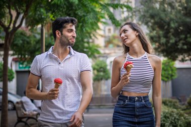 Mutlu genç çift şehirde yürüyor çilekli dondurma külahlarının tadını çıkarıyor, yaz günü gülüyor ve flört ediyorlar..