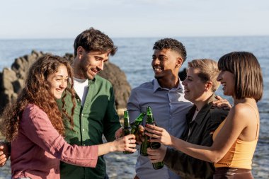 Mutlu çok kültürlü arkadaşlar deniz kenarında bira şişeleriyle kadeh kaldırarak yaz kutlamalarının ve iletişimin tadını çıkarıyorlar..