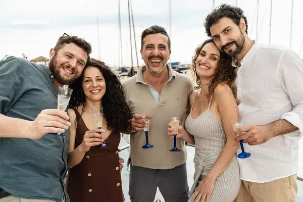 Bir yatta birbirlerine yakın duran, mavi saplı şampanya kadehleri tokuşturan ve yat limanındaki bir yaz tekne gezisinde gülen beş yetişkin arkadaştan oluşan bir grup..