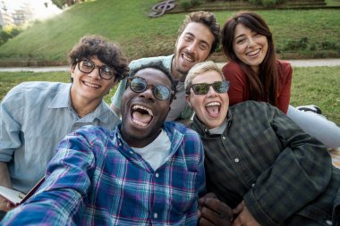 Çok ırklı Gen-Z öğrencileri, açık hava gezisi sırasında çimlerin üzerinde dinlenirken kameraya gülüyorlar ve grup selfie 'si için kollarını uzatıyorlar..