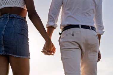 Gün batımında romantik bir yaz yürüyüşü sırasında el ele tutuşan bir çiftin yakın çekimi. Sahilde sevgi, güven ve bağlantının sembolik bir anı..
