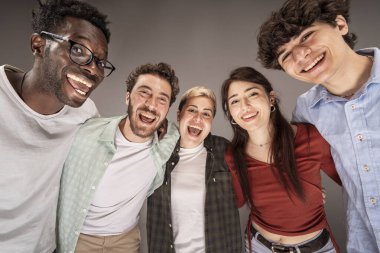 Bir grup mutlu genç arkadaş bir stüdyo ortamında birbirine yakın duruyor ve kameraya gülümsüyor, neşe, arkadaşlık ve beraberliği ifade ediyorlar..