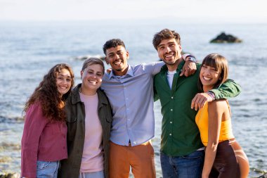 Neşeli bir grup genç arkadaş kumsalda güneşli bir günün tadını çıkarıyor, gülümsüyor ve arka planda denize sarılıyorlar..