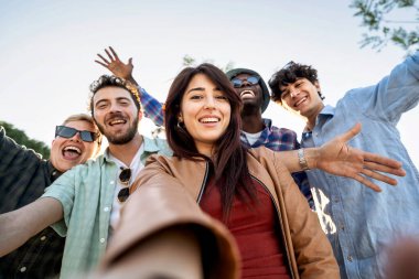 Açık hava macerası sırasında kameraya gülümseyen mutlu çoklu etnik öğrenciler, güneşli bir günde selfie için kollarını açarlar.. (