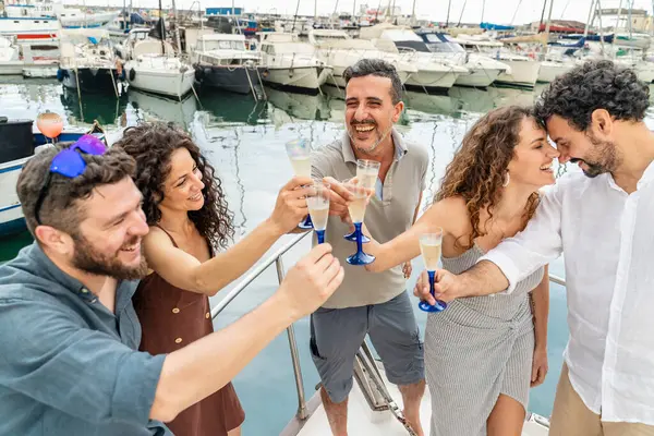 Altı yetişkin arkadaşın şampanya kadehlerini kaldırırken ve yat limanında demirlenmiş bir yatın güvertesinde kahkaha atarken yan görüntüsü. Yaz tatili kavramı, lüks yaşam tarzı ve denizde arkadaşlık..