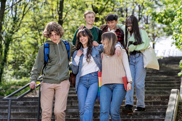 Joyful and diverse group of college students, featuring an LGBTQ rainbow pride bag strap, interacting and walking together on campus. Concepts of youth, friendship, diversity, and inclusion.