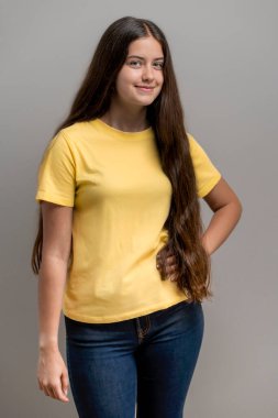 Uzun saçlı, gülümseyen genç bir kadın gri bir stüdyo arka planına karşı boş bir sarı tişört ve kot pantolon giyiyor. Kendi tasarımınız ya da markanız için çok yönlü bir model olarak mükemmel..