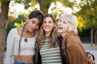 Üç mutlu genç kadından oluşan çeşitli bir grup, en iyi arkadaşlar, parkta gülüyorlar ve kucaklaşıyorlar. Gençliği, modern dostluğu, çeşitliliği ve dahil edilmeyi temsil eden gerçek bir an..