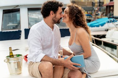 Yaz tatilinde yatta şarap ve kitapla dinlenen romantik bir çift, samimi bir liman ortamında sevgi dolu bir anı paylaşıyorlar.