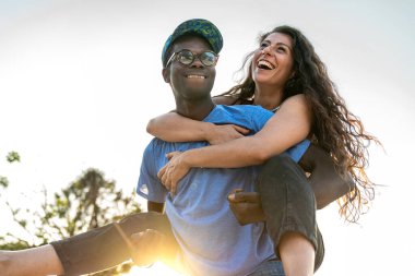 Neşeli, genç ve çeşitli bir çift gülerken adam ortağını güneşli bir günde parkta sırtında taşır. Aşk, mutluluk ve kaygısız eğlence kavramı..