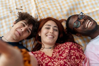A happy group of diverse young friends smile and laugh while taking a selfie, lying on a picnic blanket in a park on a sunny day.