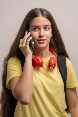 Sırt çantası ve kulaklığı olan genç bir kız öğrenci akıllı telefonuyla tarafsız bir arkaplanda konuşuyor..