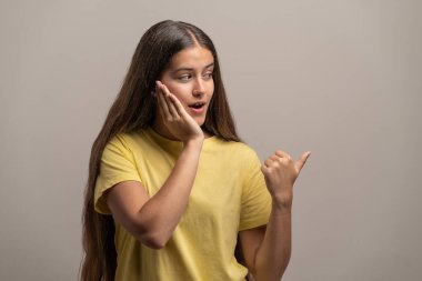 Şok olmuş bir ifadeyle genç bir kadın, elleri yanağında, inanılmaz bir şey hakkında dedikodu yaparken baş parmağıyla işaret ediyor. Söylenti, drama ve sır kavramları için mükemmel..