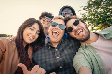 Neşeli ve çeşitli etnik çeşitlilikte genç bir arkadaş grubu kameraya gülüyor ve gülümsüyor, dışarıda eğlenceli bir selfie çekiyorlar güzel bir altın saat gün batımında.