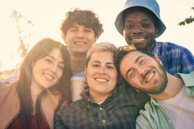 Mutlu ve çeşitli genç arkadaş grubunun otantik bir portresi, güzel bir altın saat gün batımında kameraya sımsıkı sarılıp gülümsediler..