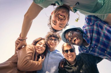 Çeşitli ve mutlu, çoklu etnik çeşitlilikte bir grup genç arkadaş, güneşli bir günde kameraya düşük bir açıdan bakıp gülerek bir çember oluşturdular..